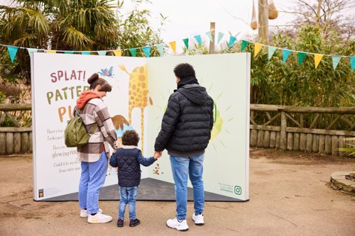 A family (a man, woman and child) look at a sculpture of a giant Poo in the Zoo book. It is taller than any of them